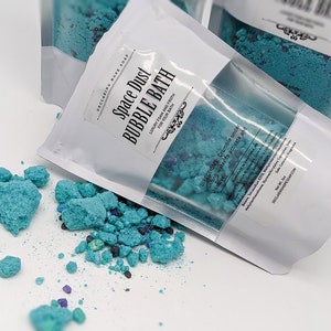 May include: White pouches of Space Dust Bubble Bath with a clear window, filled with blue bath granules. The text on the pouch reads "Space Dust Bubble Bath". Scattered granules are visible on the white surface.