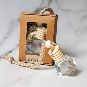 May include: A clear glass car air freshener with a wooden cap and a gold and white string. The air freshener is in a small cardboard box with a window that shows the air freshener. The box has the text "Lily of the Valley" on it.