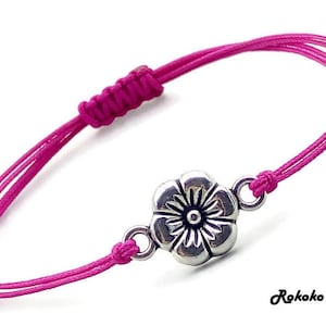 May include: A pink braided cord bracelet with a silver flower charm. The charm has a detailed design with a centre circle.