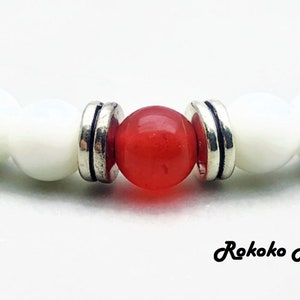 May include: A close-up of a bracelet with white and red beads. The bracelet has a brown wooden accent on each end. The red bead is in the center of the bracelet.