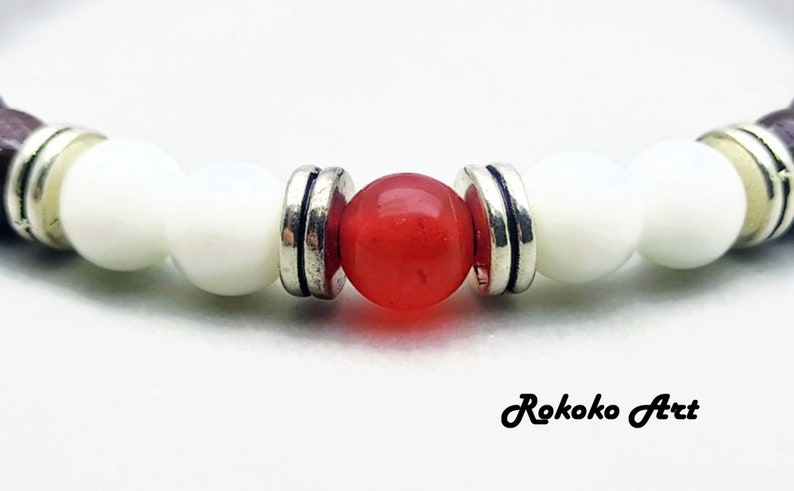 May include: A close-up of a bracelet with white and red beads. The bracelet has a brown wooden accent on each end. The red bead is in the center of the bracelet.
