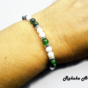May include: A white and green beaded bracelet with silver accents. The bracelet is on a person's wrist.