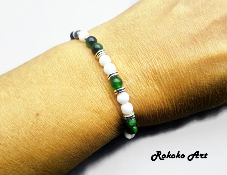 May include: A white and green beaded bracelet with silver accents. The bracelet is on a person's wrist.