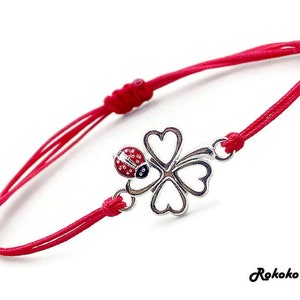 May include: Red string bracelet with a silver four-leaf clover charm and a red ladybug charm. The charm is attached to the string with a silver ring.