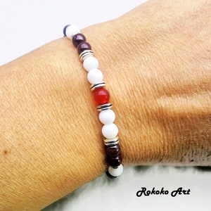 May include: A white, red, and brown beaded bracelet with silver accents. The bracelet is made with natural stones and is designed to be worn on the wrist.