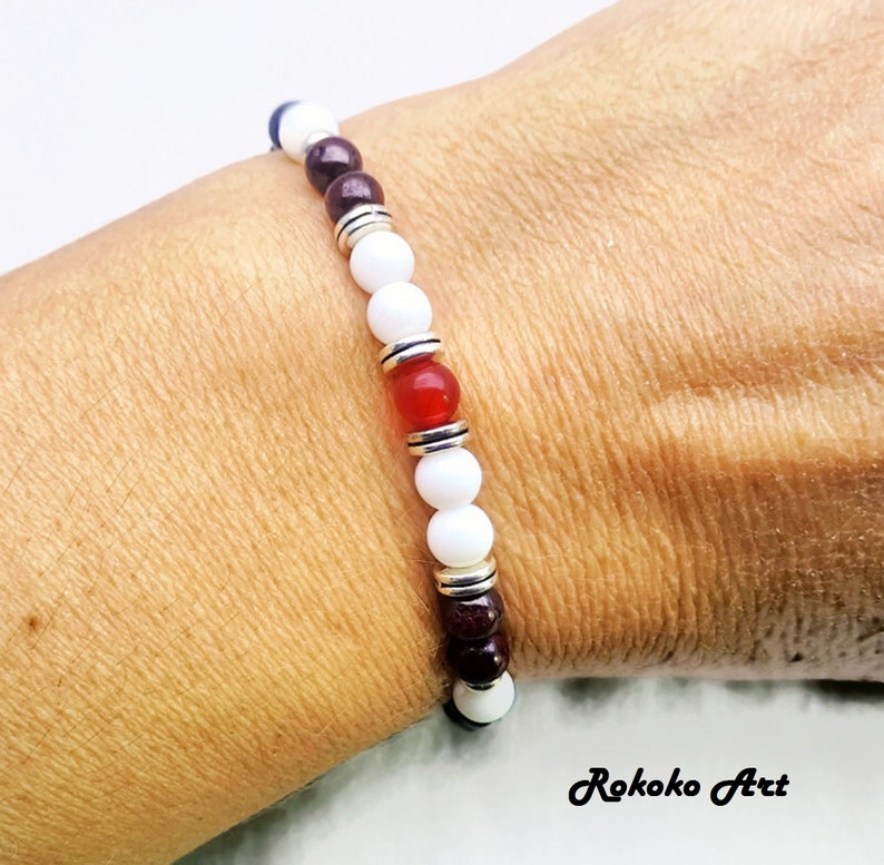 May include: A white, red, and brown beaded bracelet with silver accents. The bracelet is made with natural stones and is designed to be worn on the wrist.