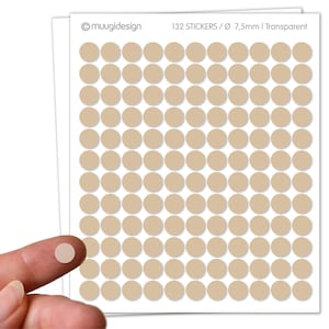 Op de afbeelding: Vel met 132 beige, transparante stickers, elk met een diameter van 7,5 mm. De stickers zijn gerangschikt in een rasterpatroon op een witte achtergrond. De tekst "muugidesign" en "132 STICKERS / Ø 7,5mm | Transparent" is afgedrukt. Een vinger houdt een sticker vast.