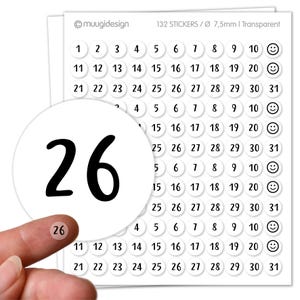 Op de afbeelding: Vel met 132 transparante stickers, elk 7,5 mm in diameter, met de nummers 1-31 en smiley's. Een vinger houdt een sticker met het nummer 26 vast. Voor kalenders of knutselwerk.