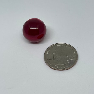 Puede incluir: Una canica de vidrio de color rojo intenso, aproximadamente del tamaño de una moneda de 25 centavos de dólar estadounidense.