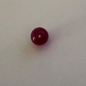 May include: A single, round, deep red bead with a small white inclusion. The bead has a glossy finish and appears to be made of a solid material. The bead is centered on a plain, off-white surface.