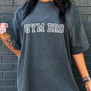 Puede incluir: Una camiseta gris oscuro de gran tamaño con el texto "GYM BRO" impreso en blanco en la parte delantera. La camiseta está hecha de un material suave y cómodo.