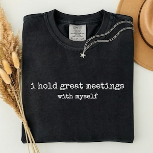May include: A black crewneck t-shirt featuring the white text "i hold great meetings with myself" printed on the chest. The shirt has a Comfort Colors brand label at the inner neckline and is displayed with a silver star pendant necklace.