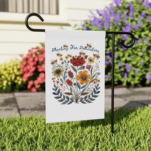 Puede incluir: Una bandera de jardín blanca con el texto "Planting For Pollinators" y un diseño floral con abejas, flores rojas, amarillas y naranjas. La bandera se muestra en un soporte de metal negro en un jardín.