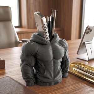 May include: A gray pen holder shaped like a muscular torso wearing a hoodie. The pen holder has a slot for pens and a phone. The desk accessory is made of a solid material and is on a wooden desk.