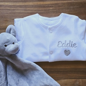 May include: A white baby onesie with a grey embroidered heart and the name "Eddie" on the front. A grey elephant plush toy with a grey blanket is in the foreground.