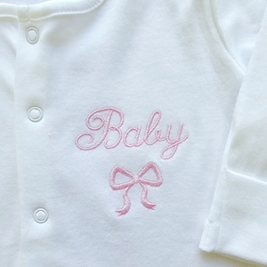 May include: White baby sleepsuit featuring the pink embroidered word "Baby" in a cursive font, and a pink bow. The sleepsuit has popper fastenings and long sleeves.