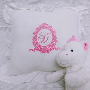 May include: White pillow with a pink embroidered monogram in a decorative frame. A white unicorn plush toy is sitting on the pillow.