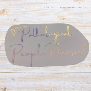 May include: A gray sticker with the words "Pathological People Pleaser" in a cursive font. The text has a holographic effect, with colors shifting from gold to purple. A heart symbol is above the word "Pathological".