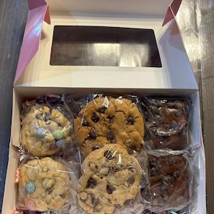 May include: A white cardboard gift box with a rectangular window containing six individually wrapped baked goods. The assortment includes two light-colored cookies with pastel candy pieces, two golden-brown chocolate chip cookies, and two dark chocolate brownies.
