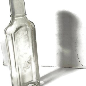 May include: A clear glass bottle with a square body and a narrow neck. The bottle is empty and has a slightly frosted appearance.