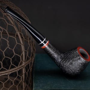 May include: A black smoking pipe with a textured bowl and a curved, glossy black stem. The pipe features a red and white accent band near the mouthpiece. The interior of the bowl has a red rim. The pipe rests on a dark green surface.