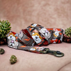 Op de afbeelding: Hondenriem met een herhalend patroon van Shiba Inu-gezichten op een grijze achtergrond. De riem heeft een rode en oranje binnenkant en een metalen sluiting. Het ontwerp bevat oranje sushirollen. Geschikt voor kleine tot middelgrote honden.