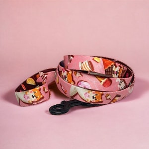 Può includere: Un guinzaglio e un collare rosa per cani con un motivo ripetuto di cani dei cartoni animati, coni gelato e altri dessert. Il guinzaglio ha un moschettone nero ed è arrotolato su uno sfondo rosa.