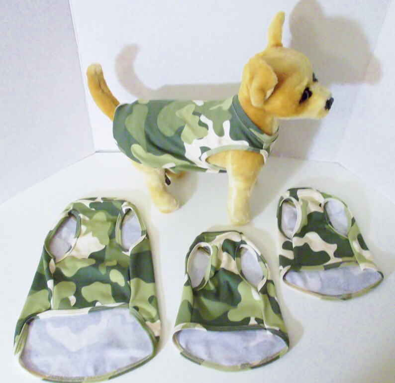 May include: Three camouflage dog shirts, one on a small dog, and two laid flat. The shirts are green and tan with a camouflage pattern.
