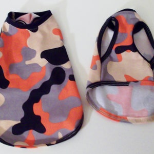 May include: Two dog shirts in a camouflage pattern with orange, black, beige, and gray colors. One shirt has a crew neck and the other has a sleeveless tank top style.