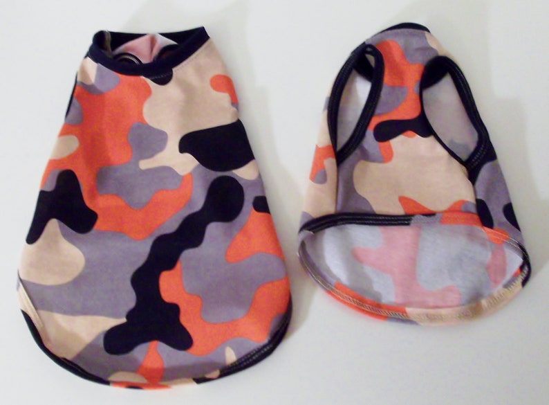 May include: Two dog shirts in a camouflage pattern with orange, black, beige, and gray colors. One shirt has a crew neck and the other has a sleeveless tank top style.