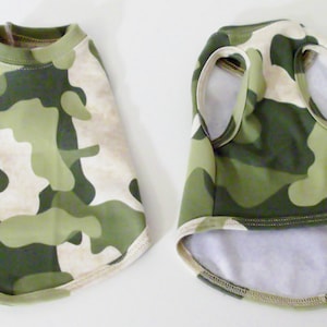 May include: Two camouflage dog shirts, one with short sleeves and one sleeveless. Both shirts are green and beige with a camouflage pattern.
