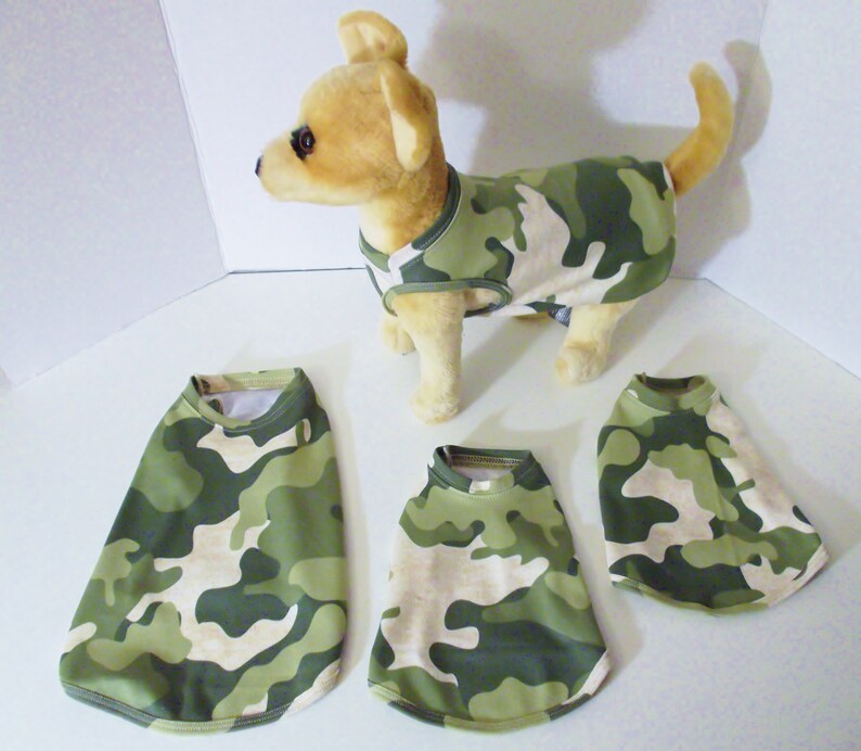 May include: Four camouflage dog shirts in green and tan. The shirts are sleeveless and have a round neckline. One shirt is being worn by a small, light brown dog.