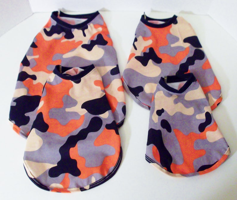 May include: Four dog shirts in different sizes, all featuring a camouflage pattern in shades of orange, black, gray, and beige.