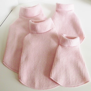 May include: Three pink turtleneck sweaters for pets, made of a ribbed knit fabric. The sweaters are different sizes, with the largest one at the top and the smallest one at the bottom.