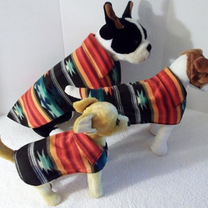 May include: Three dogs wearing colorful southwestern-style fleece dog sweaters. The sweaters have a black background with red, orange, yellow, teal, and white stripes and geometric patterns.