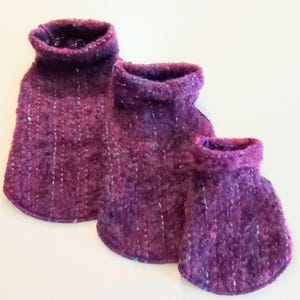 May include: Three purple tweed hot water bottle covers in varying sizes. Each cover has a rounded base and a rolled collar. The tweed fabric has a textured appearance with flecks of white and blue. The covers are arranged in a row, with the largest in the back.