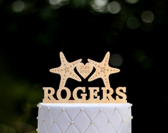 May include: A white wedding cake with a wooden starfish topper. The topper has two starfish facing each other with a heart in the middle. The heart has the date "17.04.2020" engraved on it. The cake topper also has the name "ROGERS" engraved on it.