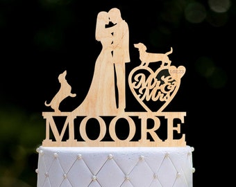Op de afbeelding: Houten taarttopper voor een bruiloft met een silhouet van een bruid en bruidegom met twee honden, een hart met de tekst "Mr & Mrs" en de datum "1.02.23", en de achternaam "MOORE".