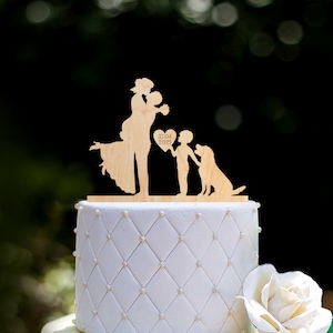 Może przedstawiać: Biały tort ślubny z drewnianym topperem przedstawiającym parę, dziecko i psa. Topper jest wygrawerowany datą "17.04.2021".