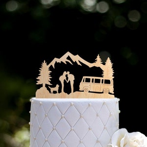 May include: Wooden wedding cake topper featuring a couple kissing in front of a mountain range, a van, and pine trees.