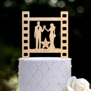 May include: Wooden wedding cake topper featuring a film strip frame with a silhouette of a couple toasting, a star with the names "Cameron & Sydney" and the date "11.12.2019".