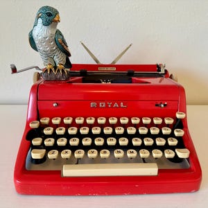 May include: A red Royal typewriter with a white keyboard and a small blue and green bird figurine on top. The typewriter has a black carriage return lever and a black space bar.