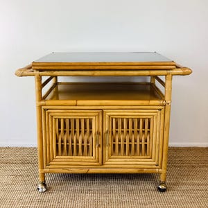 May include: A light brown bamboo bar cart with a mirrored top. The cart has two doors with woven detailing and gold-colored hardware. It sits on small wheels for easy movement. The cart has a shelf for storage.