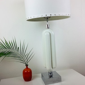 May include: White ceramic table lamp with a white fabric shade. The shade has a row of silver studs around the top edge. The lamp base is shaped like three cylinders stacked on top of each other. The base is attached to a silver square base.