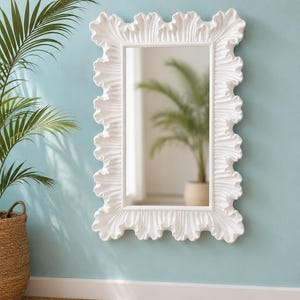 May include: A rectangular wall mirror featuring a thick, white frame with a prominent, wavy, ruffled edge design. The reflective glass surface is clear and smooth, capturing a reflection of a green plant in a pot within the room.
