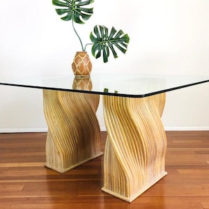 May include: A glass-top dining table with a unique, woven, natural-colored bamboo base. The base features a swirling, wave-like design. The table is set on a wooden floor.