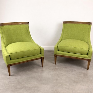 May include: Two lime green upholstered armchairs with wooden frames and legs. The chairs have a curved back and a cushioned seat. The top of the backrest has a dark wood trim. The chairs are placed against a white wall.