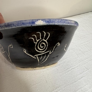 May include: A handmade ceramic bowl with a dark blue exterior and a light blue rim. The bowl features white, stylized line drawings of figures and symbols. The bowl is a unique piece of pottery.