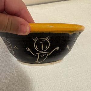 May include: A small, handmade ceramic bowl with a black exterior and a bright yellow rim. The bowl features white, whimsical line drawings of figures and shapes. The bowl is held by a hand, showcasing its size and design.