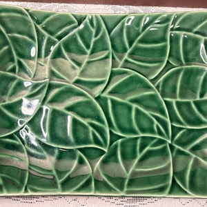 May include: A rectangular ceramic tray featuring a glossy, deep green glaze with a raised, embossed pattern of overlapping leaves. The surface has a smooth, reflective finish with subtle variations in color depth across the leaf veins.